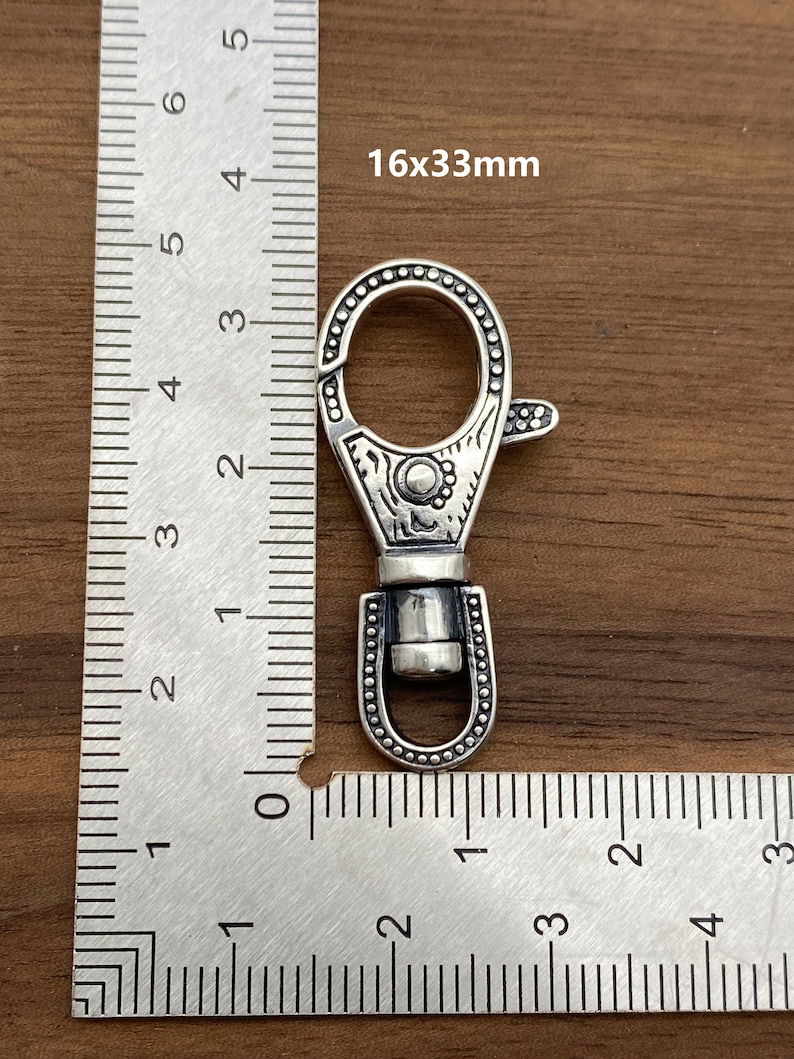 May include: A silver-colored swivel clasp with an ornate design, measuring 16x33mm. The clasp features a decorative pattern around the edges and a spring-loaded lever for secure closure. The image includes a ruler for scale.