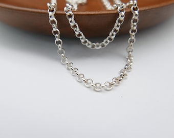 May include: A silver-colored necklace with a delicate chain design. The necklace is composed of small, circular links, creating a simple yet elegant aesthetic. The chain is displayed against a white surface and a brown wooden bowl.