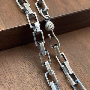 Puede incluir: Un collar de cadena de plata con un diseño de eslabón cuadrado. La cadena tiene un cierre de mosquetón.