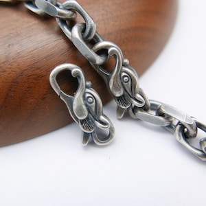 May include: Close-up of a silver chain necklace with a unique design. The chain links are connected with a clasp featuring swan-like heads. The necklace is displayed on a wooden surface.