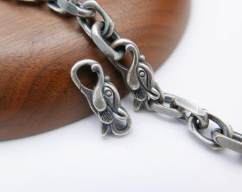 May include: Close-up of a silver chain necklace with a unique design. The chain links are connected with a clasp featuring swan-like heads. The necklace is displayed on a wooden surface.