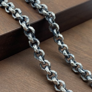 Puede incluir: Primer plano de una cadena de plata con un diseño texturizado y martillado. Los eslabones de la cadena son de forma ovalada y entrelazados, creando un aspecto audaz e industrial. La cadena se muestra sobre una superficie de madera.