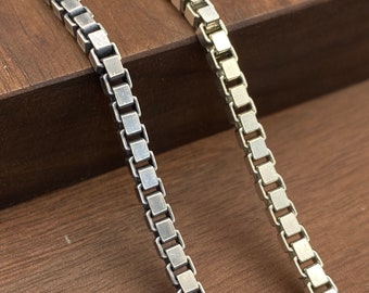 Op de afbeelding: Twee zilveren kettingen met vierkante schakels, de ene ketting is iets dikker dan de andere, beide kettingen liggen op een houten oppervlak.