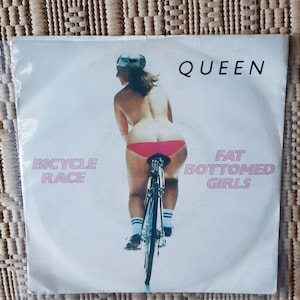 Könnte beinhalten: Eine weiße 7-Zoll-Vinyl-Schallplatte mit einem rosa und weißen Etikett. Das Etikett zeigt eine Frau, die Fahrrad fährt, mit dem Text "Bicycle Race" und "Fat Bottomed Girls". Die Schallplatte ist von der Band Queen.