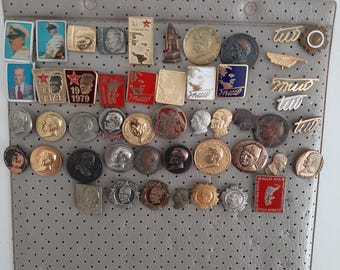 Op de afbeelding: Een verzameling vintage emaille-speldjes met verschillende ontwerpen, waaronder portretten, symbolen en tekst. De speldjes worden gepresenteerd op een grijze achtergrond met een rasterpatroon.