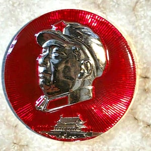 May include: A circular metal pin featuring a silver-toned profile relief of a person wearing a cap with a star, set against a vibrant red background with a sunburst pattern. At the bottom, a silver-toned relief of a traditional building is visible.
