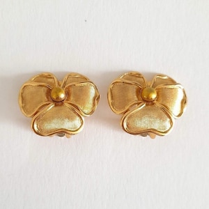 Könnte beinhalten: Ein Paar goldfarbene Metall-Clip-Ohrringe in Form von dreiblättrigen Blumen. Jeder Ohrring verfügt über eine texturierte, gebürstete Goldoberfläche auf den Blütenblättern und einen glatten, polierten goldfarbenen Mittelpunkt.