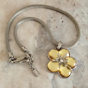 May include: A silver-toned mesh chain necklace featuring a large, five-petaled flower pendant. The flower has a smooth, yellow-gold colored center with a silver-toned stamen detail accented by small, clear stones. A small rectangular tag is attached.