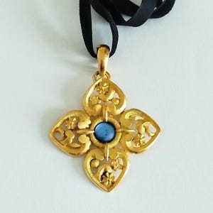 Könnte beinhalten: Ein goldfarbener Anhänger mit einem komplizierten, durchbrochenen Blumen- und Herzmuster und einem runden, blauen Cabochon-Stein in der Mitte. Der Anhänger hängt an einem schwarzen Band, was eine Halskette mit matter, strukturierter Oberfläche ergibt.