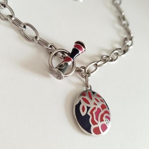 May include: A silver-toned metal chain necklace featuring a toggle clasp with a black, red, and white enamel finish. An oval-shaped pendant hangs from the clasp, decorated with a floral-inspired pattern in shades of red, pink, and dark blue enamel.