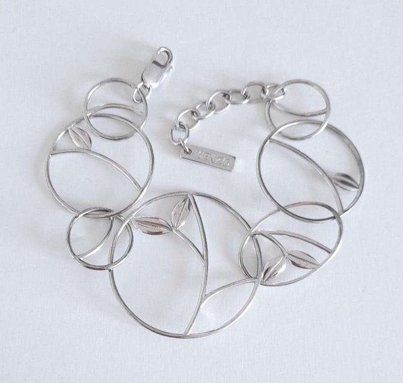 K&ouml;nnte beinhalten: Ein silberfarbenes Metallarmband bestehend aus einer Reihe miteinander verbundener kreisf&ouml;rmiger Glieder. Mehrere Kreise enthalten zarte, blattf&ouml;rmige Ausschnitte. Das Armband verf&uuml;gt &uuml;ber einen Karabinerverschluss und ein kleines Schild mit der Gravur KENZO.