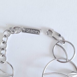 K&ouml;nnte beinhalten: Ein silberfarbenes Metallkettenarmband mit kreisf&ouml;rmigen Gliedern und einem rechteckigen Anh&auml;nger, der mit dem Text KENZO graviert ist. Das Armband verf&uuml;gt &uuml;ber einen Karabinerverschluss und eine Reihe miteinander verbundener Ringe.