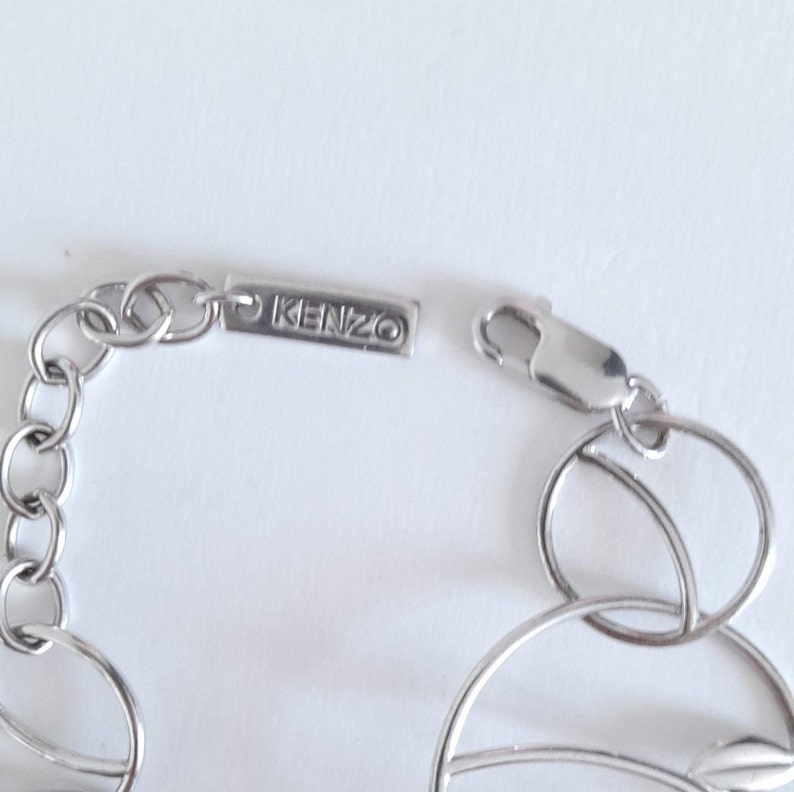 K&ouml;nnte beinhalten: Ein silberfarbenes Metallkettenarmband mit kreisf&ouml;rmigen Gliedern und einem rechteckigen Anh&auml;nger, der mit dem Text KENZO graviert ist. Das Armband verf&uuml;gt &uuml;ber einen Karabinerverschluss und eine Reihe miteinander verbundener Ringe.