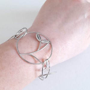 K&ouml;nnte beinhalten: Ein silberfarbenes Armband mit einer Reihe von miteinander verbundenen kreisf&ouml;rmigen Gliedern. Jedes kreisf&ouml;rmige Glied enth&auml;lt einen zarten, d&uuml;nnen Metallstiel mit zwei kleinen, glatten blattf&ouml;rmigen Akzenten am inneren Rand des Rahmens.