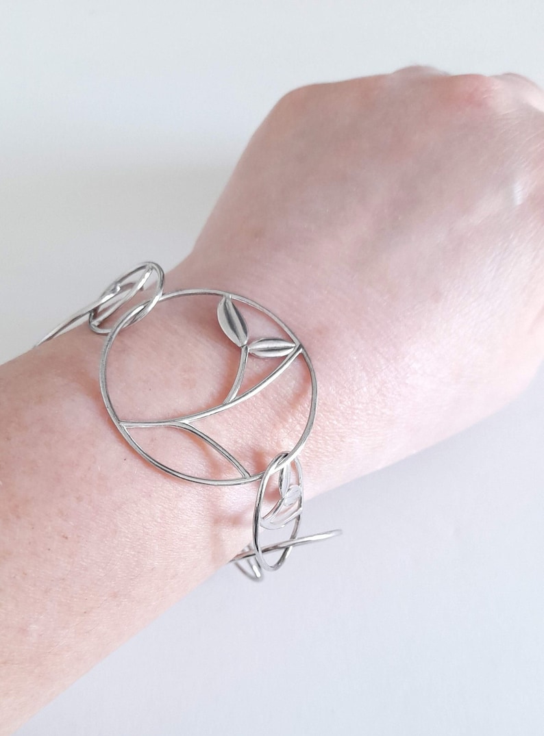 K&ouml;nnte beinhalten: Ein silberfarbenes Armband mit einer Reihe von miteinander verbundenen kreisf&ouml;rmigen Gliedern. Jedes kreisf&ouml;rmige Glied enth&auml;lt einen zarten, d&uuml;nnen Metallstiel mit zwei kleinen, glatten blattf&ouml;rmigen Akzenten am inneren Rand des Rahmens.