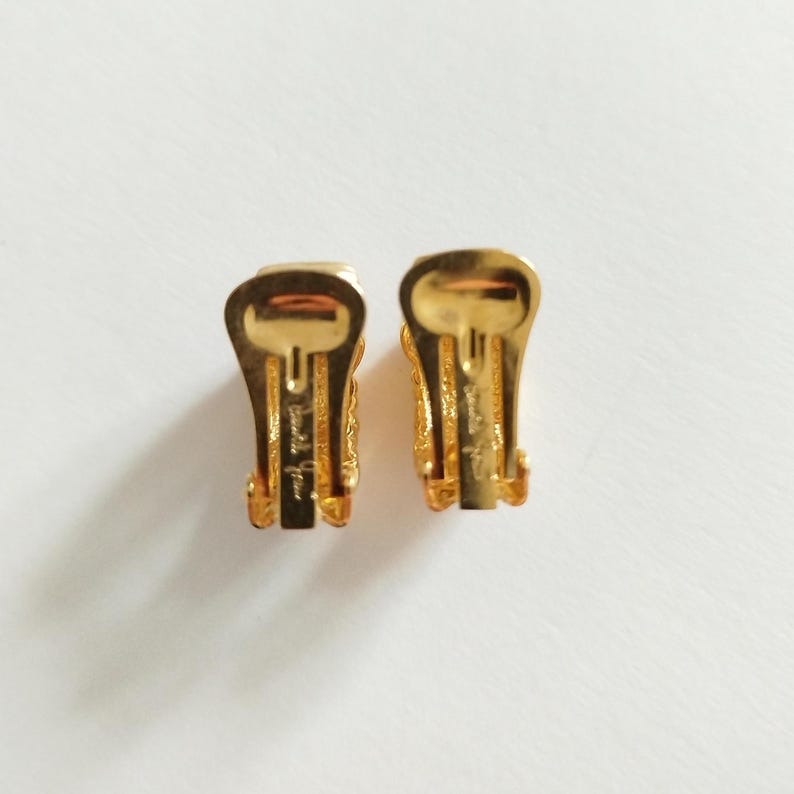 K&ouml;nnte beinhalten: Ein Paar goldfarbene Ohrclips mit einem detaillierten Design. Die Ohrringe sind auf der R&uuml;ckseite mit "Judith Leiber New York" gekennzeichnet.