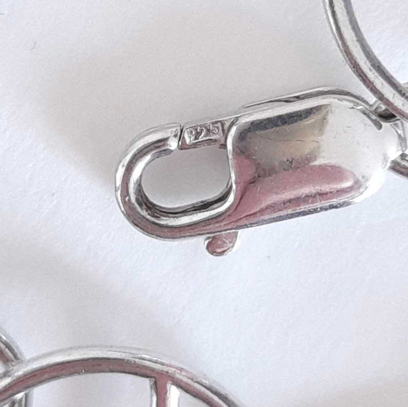 K&ouml;nnte beinhalten: Ein silberfarbener Karabinerverschluss aus Metall von einer Schmuckkette. Der Verschluss weist einen kleinen, rechteckigen Stempel auf, der deutlich die Zahl 925 zeigt und die Reinheit des Metalls angibt. Der Mechanismus ist in geschlossener Position.