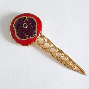 Peut inclure: Une broche en métal doré avec une tête circulaire bordée d'émail rouge vif entourant un centre ornemental violet foncé aux motifs floraux. La tige de la broche est allongée et composée d'une série de cercles dorés interconnectés.