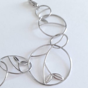 K&ouml;nnte beinhalten: Ein silberfarbenes Metallarmband bestehend aus miteinander verbundenen kreisf&ouml;rmigen Gliedern. Jedes kreisf&ouml;rmige Glied weist ein zartes internes Zweigdesign mit kleinen, glatten, blattf&ouml;rmigen Akzenten auf. Das Armband hat einen Karabinerverschluss.