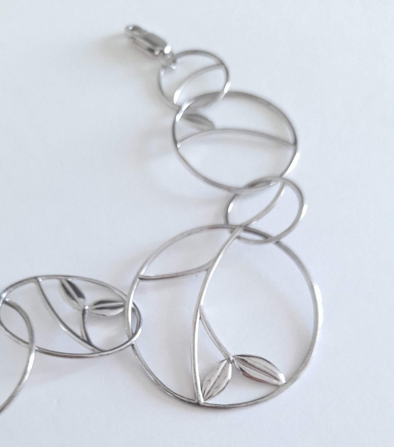 K&ouml;nnte beinhalten: Ein silberfarbenes Metallarmband bestehend aus miteinander verbundenen kreisf&ouml;rmigen Gliedern. Jedes kreisf&ouml;rmige Glied weist ein zartes internes Zweigdesign mit kleinen, glatten, blattf&ouml;rmigen Akzenten auf. Das Armband hat einen Karabinerverschluss.