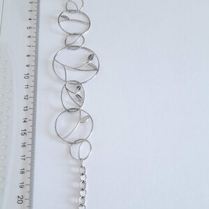 K&ouml;nnte beinhalten: Ein silberfarbenes Metallarmband mit einer Reihe von miteinander verbundenen kreisf&ouml;rmigen Gliedern, von denen einige blattartige Motive enthalten. Ein durchsichtiges Kunststofflineal daneben zeigt eine Gesamtl&auml;nge von 22 Zentimetern.