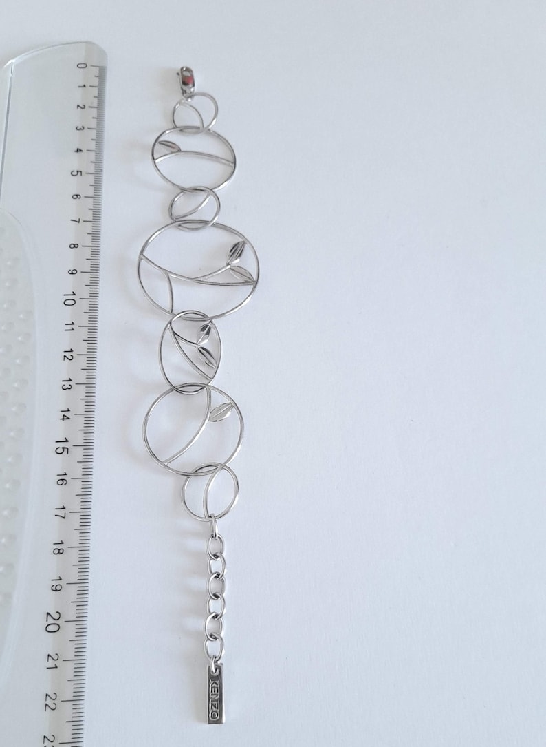 K&ouml;nnte beinhalten: Ein silberfarbenes Metallarmband mit einer Reihe von miteinander verbundenen kreisf&ouml;rmigen Gliedern, von denen einige blattartige Motive enthalten. Ein durchsichtiges Kunststofflineal daneben zeigt eine Gesamtl&auml;nge von 22 Zentimetern.