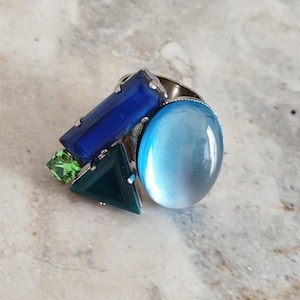 Op de afbeelding: Een statement ring met een grote ovale lichtblauwe cabochon, een rechthoekige diepblauwe steen, een donkergroene driehoekige steen en een kleine vierkante limoengroene steen, gezet in een zilverkleurige metalen basis met zichtbare zettingen.