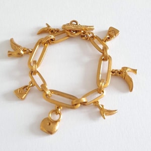 Puede incluir: Una pulsera de dijes en tono dorado que presenta una serie de eslabones ovalados alargados conectados por un cierre de barra. La pulsera está adornada con múltiples dijes pequeños, incluyendo varias formas de zapatos de tacón, un bolso y un candado en forma de corazón.