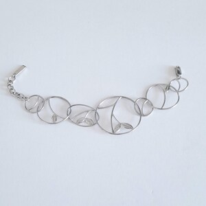 K&ouml;nnte beinhalten: Ein silberfarbenes Metallarmband mit einer Reihe von miteinander verbundenen kreisf&ouml;rmigen Gliedern unterschiedlicher Gr&ouml;&szlig;e. Mehrere der kreisf&ouml;rmigen Rahmen enthalten zarte, blattf&ouml;rmige Ausschnitte oder interne Drahtdetails.
