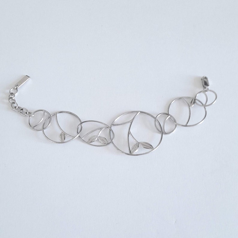 K&ouml;nnte beinhalten: Ein silberfarbenes Metallarmband mit einer Reihe von miteinander verbundenen kreisf&ouml;rmigen Gliedern unterschiedlicher Gr&ouml;&szlig;e. Mehrere der kreisf&ouml;rmigen Rahmen enthalten zarte, blattf&ouml;rmige Ausschnitte oder interne Drahtdetails.