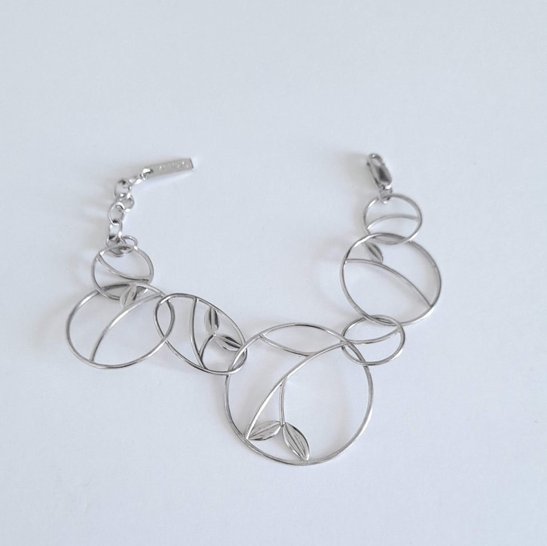 K&ouml;nnte beinhalten: Ein silberfarbenes Metallarmband mit einer Reihe von miteinander verbundenen kreisf&ouml;rmigen und ovalen Gliedern. Mehrere dieser offenen Glieder enthalten kleine, blattf&ouml;rmige Metallakzente. Das Armband verf&uuml;gt &uuml;ber eine kurze Verl&auml;ngerungskette.