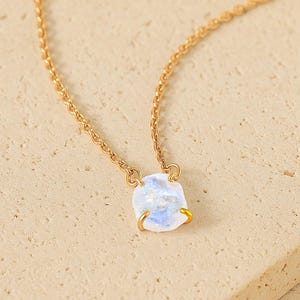 May include: A gold-colored necklace with a small, square, light blue and white gemstone pendant. The necklace chain is delicate and the pendant is set in a gold-colored bezel. The necklace is displayed on a light tan surface.