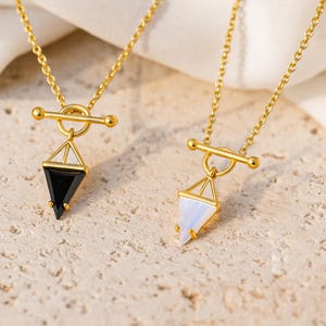 May include: Two gold-colored necklaces with geometric pendants. One pendant is a black triangle, the other is a white triangle. The necklaces have a bar and ring clasp. The background is a light beige, textured surface.