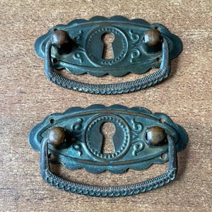 May include: Two antique, oval-shaped, metal drawer pulls with ornate designs. Each pull has a keyhole cutout and a rounded knob on each side. The pulls are a dark green color with a distressed finish.