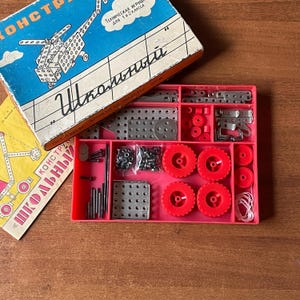 May include: A vintage metal construction set with a blue and orange box featuring a helicopter illustration. The set includes various metal pieces, red wheels, and a pink tray. The box reads "School" in Russian.