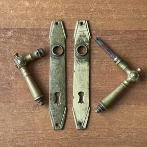 Pode incluir: Conjunto de ferragens de porta antigas em latão, incluindo duas placas longas com buracos de fechadura e duas maçanetas de alavanca. As ferragens têm uma aparência vintage e envelhecida, com uma cor castanho-dourada quente. O conjunto está disposto sobre uma superfície de madeira.