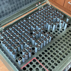 May include: A metal storage case filled with numerous cylindrical objects. Each object has a black top with white text and is arranged in a grid pattern. The case is open, revealing the contents, and has a green exterior.