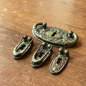 May include: Antique brass hardware set featuring an oval escutcheon with a keyhole and handle, along with three matching pulls. The pieces have ornate floral detailing and a vintage aesthetic. The hardware is likely for a cabinet or drawer.