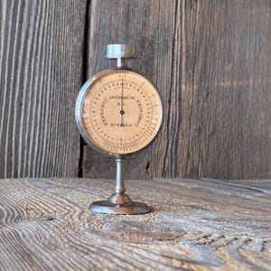May include: A vintage metal spherometer with a circular beige dial featuring black markings and text that reads SPHÉROMÈTRE G.L. CONVEXES CONCAVES B.S.D.G. The instrument has a central needle, a silver-toned metallic frame, and a circular base stand.