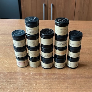 May include: A collection of vintage black film canisters with black lids and tan labels. The labels feature Cyrillic text. The canisters are stacked on a wooden surface.