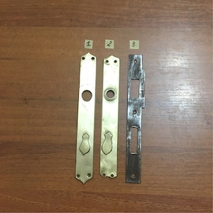 May include: Three metal door hardware pieces, two are brass and one is steel. The brass pieces are rectangular with a keyhole and a decorative design at the bottom. The steel piece is rectangular with a keyhole and a rectangular cutout in the middle.