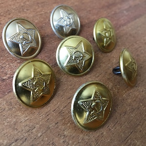 Może przedstawiać: Osiem złotych metalowych guzików z pięcioramienną gwiazdą i symbolem młota i sierpa w środku. Guziki mają podniesiony brzeg i gładką powierzchnię.