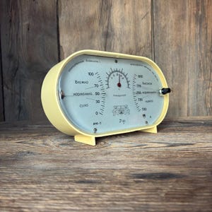 Puede incluir: Una estación meteorológica ovalada vintage con carcasa de plástico amarillo pálido y frente de cristal. La esfera muestra texto en cirílico, un termómetro central, un higrómetro a la izquierda y un barómetro a la derecha con un mando.