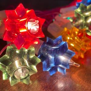 Puede incluir: Primer plano de una cadena de luces navideñas con bombillas en forma de estrella en rojo, verde, azul y amarillo. Las bombillas son de plástico translúcido y tienen un diseño retro. Las luces están sobre una superficie marrón.