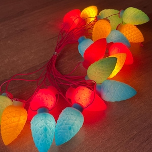 Pode incluir: Uma sequência de luzes de Natal retrô em forma de abacaxi, em várias cores. As luzes incluem vermelho, amarelo, verde e azul, e são conectadas por um fio vermelho. Uma decoração nostálgica e festiva para a época natalícia.