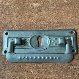 Puede incluir: Un tirador de cajón de metal plateado con una cerradura y detalles decorativos. El tirador tiene una forma rectangular con bordes redondeados y un asa elevada.