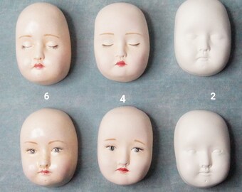 May include: Six porcelain doll heads, some with painted features, arranged in rows of three. The heads are all white and have a smooth, matte finish. The heads with painted features have closed eyes, rosy cheeks, and red lips.