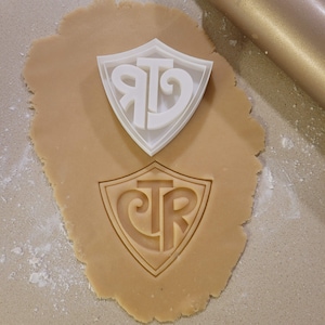 Op de afbeelding: Een witte plastic koekjesvormer in de vorm van een schild met de letters "CR" erin. De koekjesvormer ligt op een rond stuk koekdeeg met hetzelfde ontwerp dat in het deeg is gedrukt.