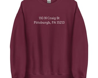 May include: A maroon sweatshirt with the text "110 N Craig St Pittsburgh, PA 15213" printed in white. The sweatshirt is a crew neck style and appears to be made of a soft material.
