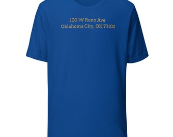 May include: Royal blue t-shirt with short sleeves. The shirt features the text "100 W Reno Ave Oklahoma City, OK 73102" printed in a gold color. The shirt is made of a soft material.